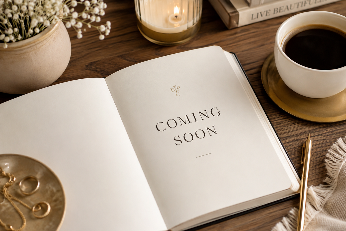 Open notebook on a wooden table with “Coming Soon” written on the page, styled with coffee, candle, flowers, and a gold pen.